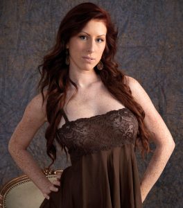 Red headed woman wearing lacy brown nighties against a light grey background. Her dark red hair spills over her shoulders and slightly covers her bare arms.
